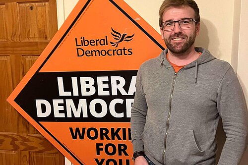 candidate, Pele Barnes, standing in front of a stakeboard saying Liberal Democrats, Working For You.