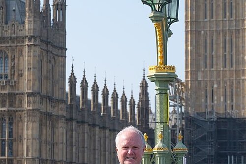 Clive Jones MP in a suit with a yellow tie. He is stood on Westminster Bridge with the palace of Westminster in the background. He is smiling at the camera.